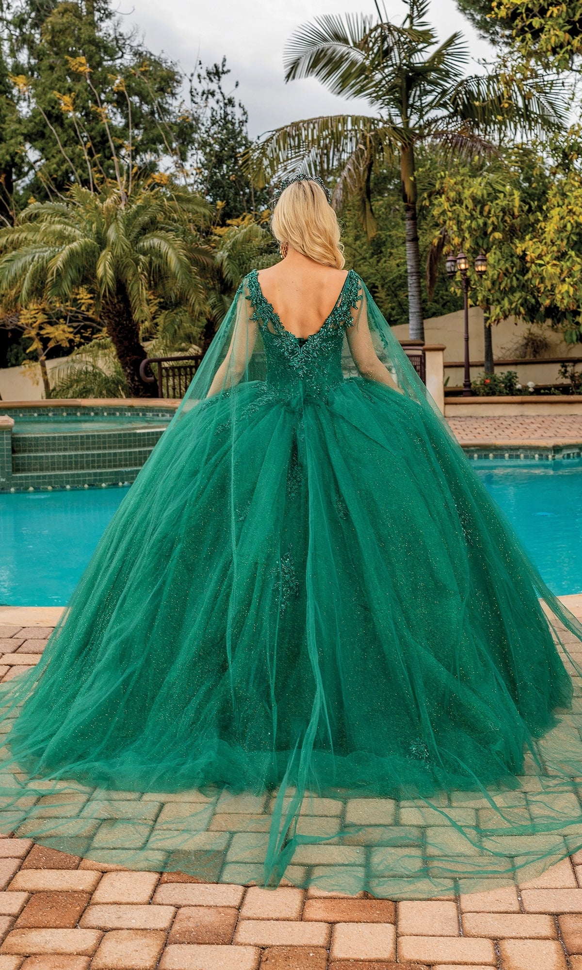 Wearing the Quinceanera Dress 1767 By Dancing Queen, a person in a green, floor-length ball gown with a full tulle skirt and sheer cape stands poolside, facing away toward a garden with palm trees.
